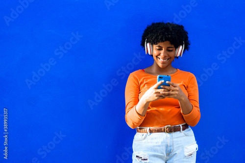 Happy beautiful afro american woman with orange sweater and white headphones listening to music on a phone, music and technology concept.