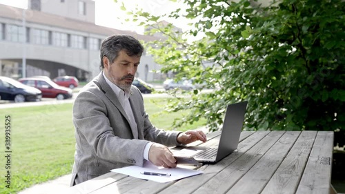 An adult businessman wearing an American jacket works outdoors in a park with buildings