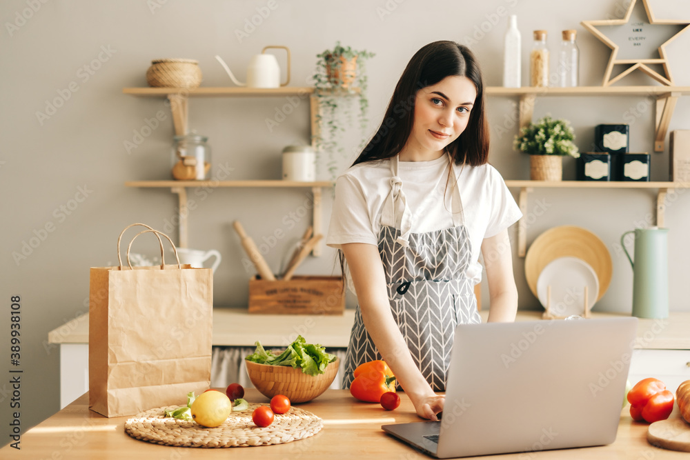 Young caucasian woman in apron use laptop computer in the modern kitchen, preparing salad, read recipe. Young caucasian woman in apron use laptop computer in the modern kitchen, preparing salad, read recipe.