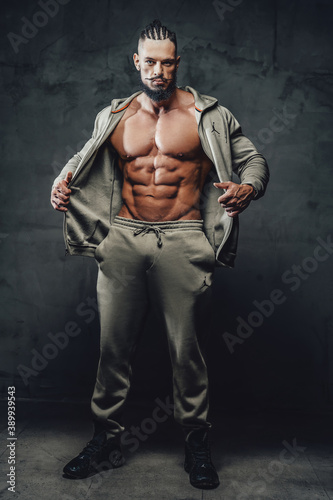 Handsome and powerful athlete with naked torso and perfect abs and with beard and moustache poses in dark background.