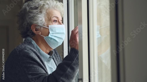 Senior woman with face mask stay at home during Covid-19 pandemic. Depressed old aged woman looking outside of a window with surgical mask during quarantine and lockdown measures for Covid19. photo