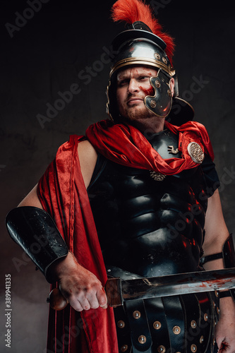 Strong and warlike legionary with blood on his face in black armour with red cape poses holding a sword and looking away.