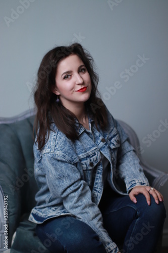 attractive brunette in jeans and denim jacket sitting on grey chair. studio shoot. 