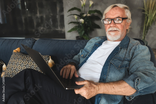 Φωτογραφία Elderly man with glasses with gray hair and a beard sits at home on the sofa in