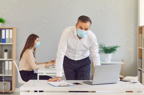 Adult man in a protective medical mask stands in the office against the background of his colleague.