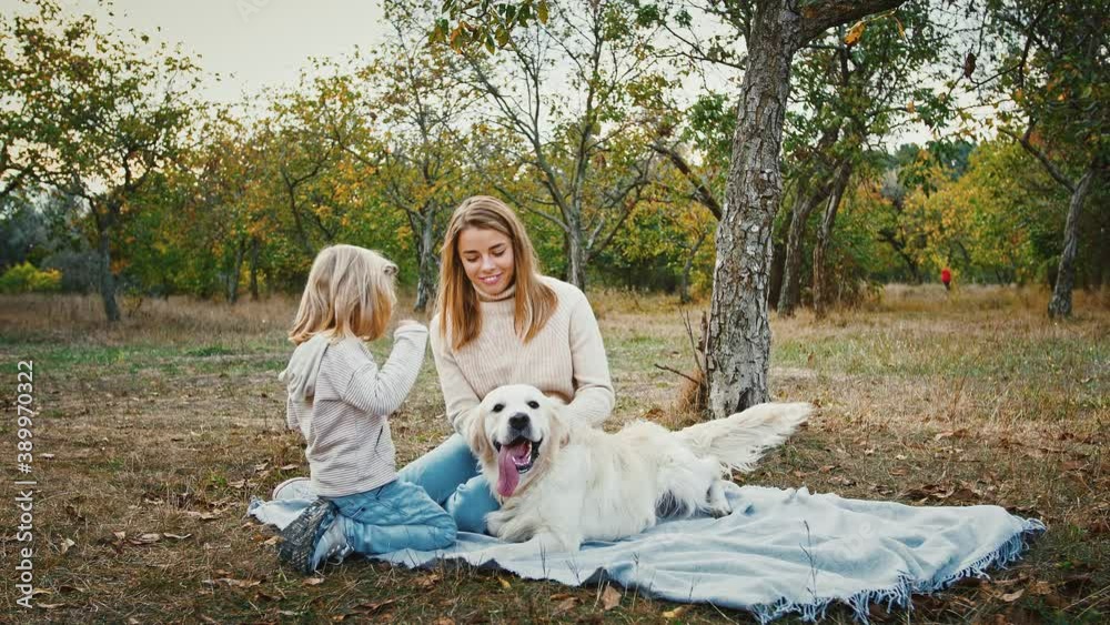 Young mum and little daughter sitting on picnic blanket, smiling and stroking their dog during walk in autumn park Young mum and little daughter sitting on picnic blanket, smiling and stroking their dog during walk in autumn park