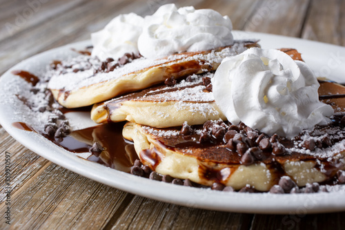 A closeup view of a plate of chocolate chip pancakes, featuring whipped cream and powdered sugar toppings, in a restaurant or kitchen setting.