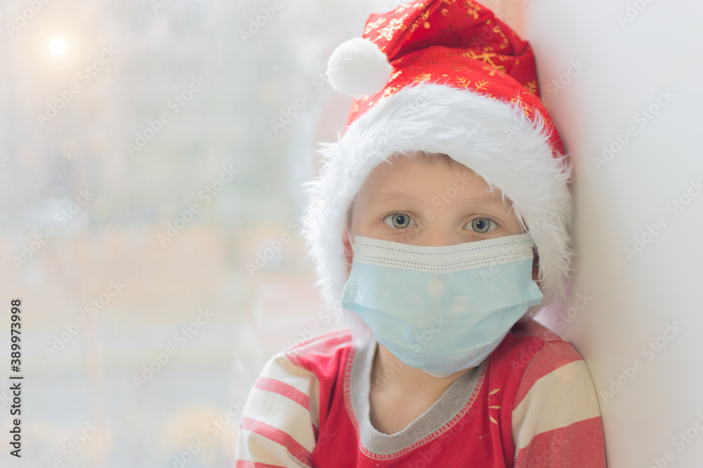 preschooler in a medical mask and a New Year's cap looks at the camera against the background of a window. Waiting for Christmas and New Year in modern reality and the covid-19 pandemic. Soft focus. preschooler in a medical mask and a New Year's cap looks at the camera against the background of a window. Waiting for Christmas and New Year in modern reality and the covid-19 pandemic. Soft focus.