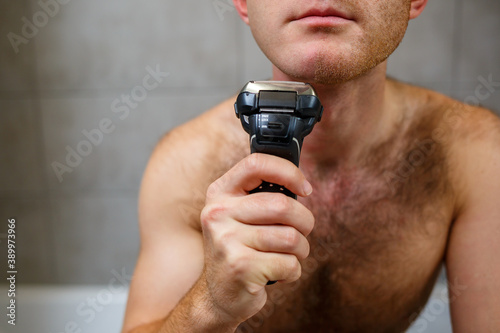 A man shaves his face with an electric razor in front of a mirror. Skin irritation. Bath procedure
