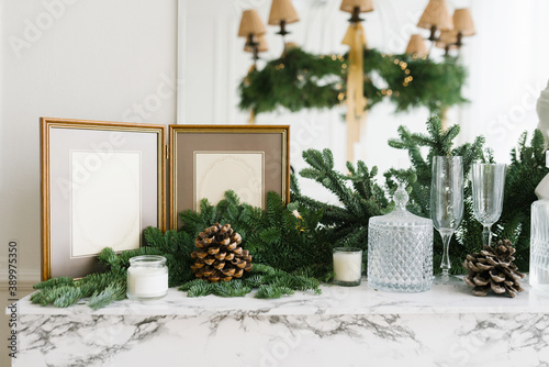 Christmas decor in home decoration: photo frame, spruce branches, glasses, candles and cones on a marble shelf