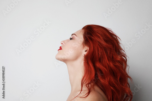 Side headshot of young woman with beautiful long red hair posing over white wall