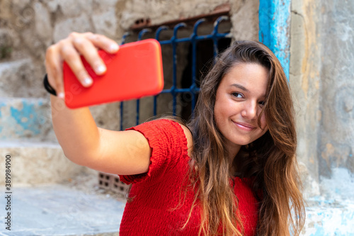 attractive girl dressed in red dress taking a selfie in the street on a sunny day and smiling © Supermelon