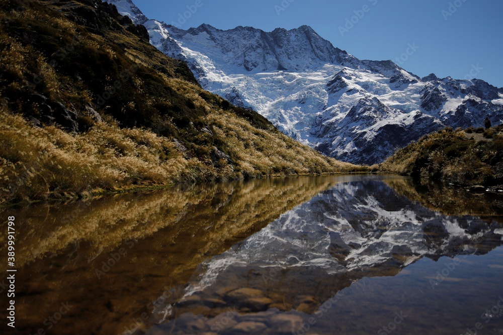 Symmetrical picture of a snow covered mountain and yellow grasses reflecting in a lake at the Sealy Tarns viewpoint, Canterburry, New Zealand