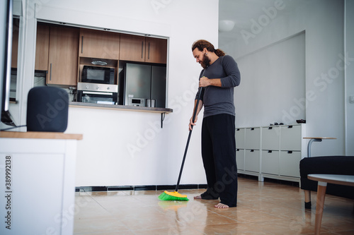 A bearded man wearing a pajama uses a sweep to broom the floor of his living room