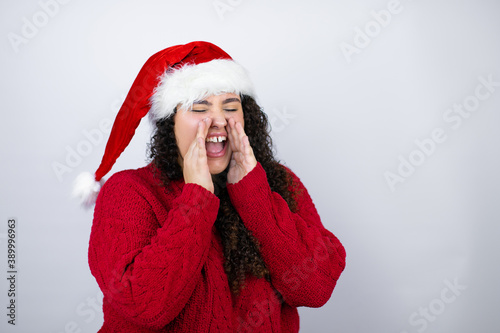 Young beautiful woman wearing a Santa hat over white background shouting and screaming loud to side with hands on mouth