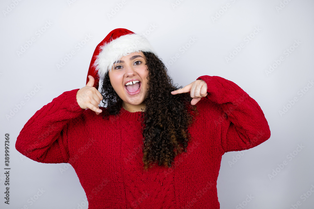 Young beautiful woman wearing a Santa hat over white background doing the “call me” gesture with her hands. Young beautiful woman wearing a Santa hat over white background doing the “call me” gesture with her hands.