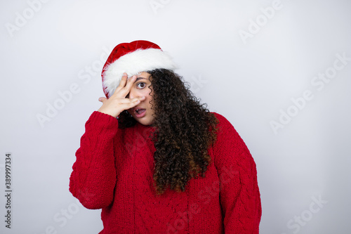 Young beautiful woman wearing a Santa hat over white background peeking in shock covering face and eyes with hand  looking through fingers with embarrassed expression