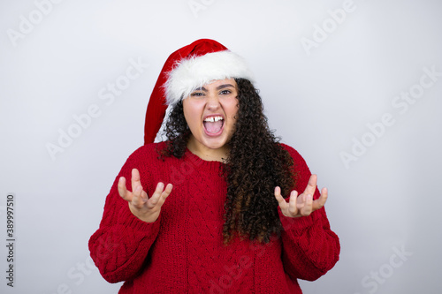 Young beautiful woman wearing a Santa hat over white background crazy and mad shouting and yelling with aggressive expression and arms raised. Frustration concept.