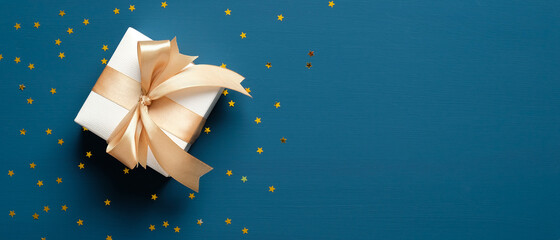 White gift box with golden ribbon bow on blue background with confetti stars. Christmas present, valentine day surprise, birthday concept. Flat lay, top view, copy space.