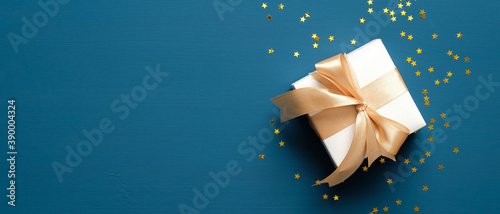 White gift box with golden ribbon bow on blue background with confetti. Christmas present, valentine day surprise, birthday concept. Flat lay, top view. photo