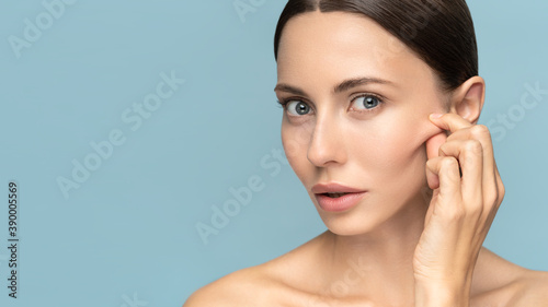 Woman without makeup touching cheeks after glycolic acid peel, has signs of aging skin on her face, looking at camera, isolated on studio blue background. Beauty skincare, cosmetology facial treatment