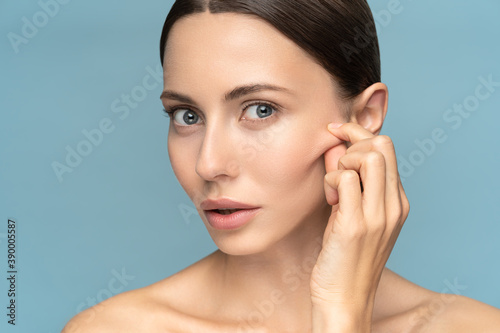Woman without makeup touching cheeks after glycolic acid peel, has signs of aging skin on her face, looking at camera, isolated on studio blue background. Beauty skincare, cosmetology facial treatment photo