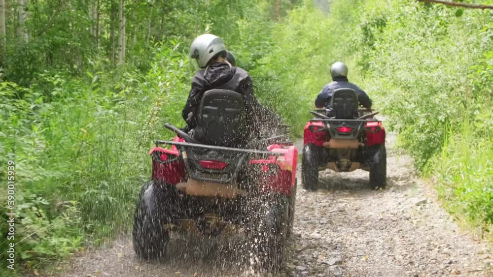 Tracking shot of people in helmets riding quad bikes through puddles along narrow forest road in rough terrain Tracking shot of people in helmets riding quad bikes through puddles along narrow forest road in rough terrain