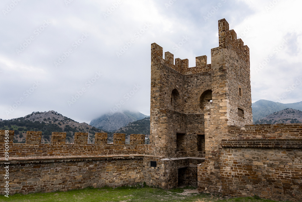 Tower and wall of Genoese fortress of 14th century in the Sudak bay on the Peninsula of Crimea on the background of mountains and cloudy sky