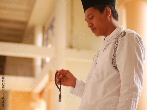 Indonesian muslim young man wearing medical face mask standing while raised hands and praying. Indonesiam moslem man praying with the prayer beads in his hand. photo