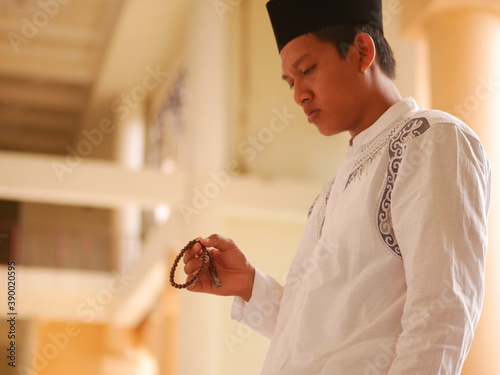 Indonesian muslim young man wearing medical face mask standing while raised hands and praying. Indonesiam moslem man praying with the prayer beads in his hand. photo