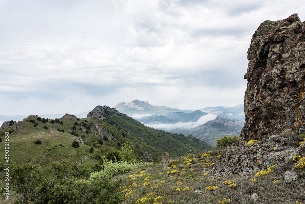 The slope of the mountain with yellow flowers and a vertical stone column against the mountains in the clouds