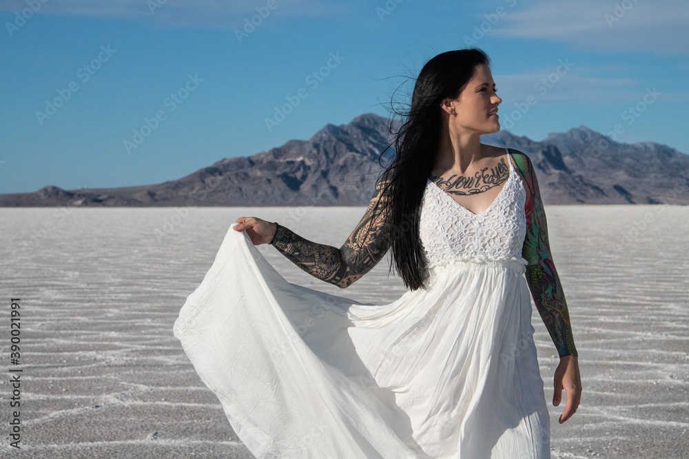 Young woman in a white dress holding one side of the dress. Woman has brown hair and tattoo sleeves. She is looking in the distance and appears happy Young woman in a white dress holding one side of the dress. Woman has brown hair and tattoo sleeves. She is looking in the distance and appears happy