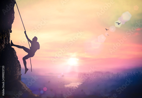 Adventure concept: Silhouette bold heroic man try to climb with rope over natural rocky wall wide valley autumn sunset mountain background