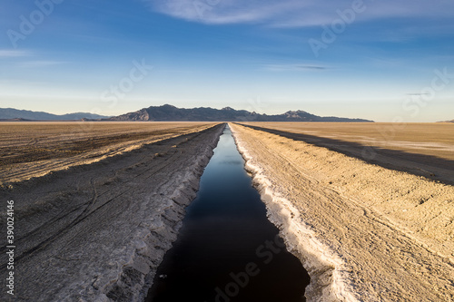 Aerial view of a straight river in the middle of the Bonneville Salt Flats in Utah. Mountains can be seen in the distance and the stream is blue compared with the white of the surrounding salt.