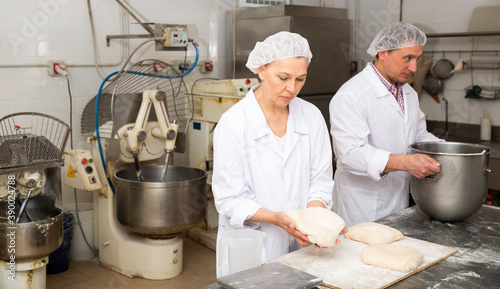 Mature woman professional baker standing at work table and forming dough for baking bread