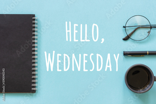 Hello Wednesday words in black notepad, pen, cup of coffee and reeding glasses on blue background. Concept Happy Wednsday. Top view Flat lay