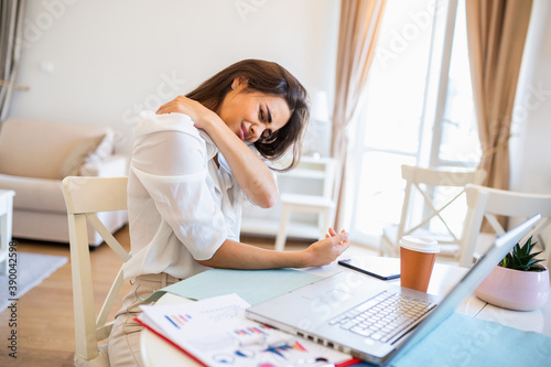 Portrait of young stressed woman sitting at home office desk in front of laptop, touching aching back with pained expression, suffering from backache after working on laptop photo