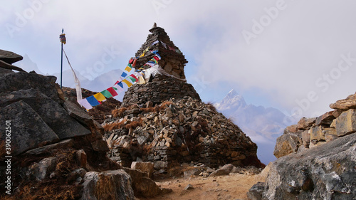 Buddhist stone monument with attached colorful prayer flags besides hiking trail near Phortse, Sagarmatha National Park, Himalayas, Nepal with majestic mountains vanishing in the rising clouds.