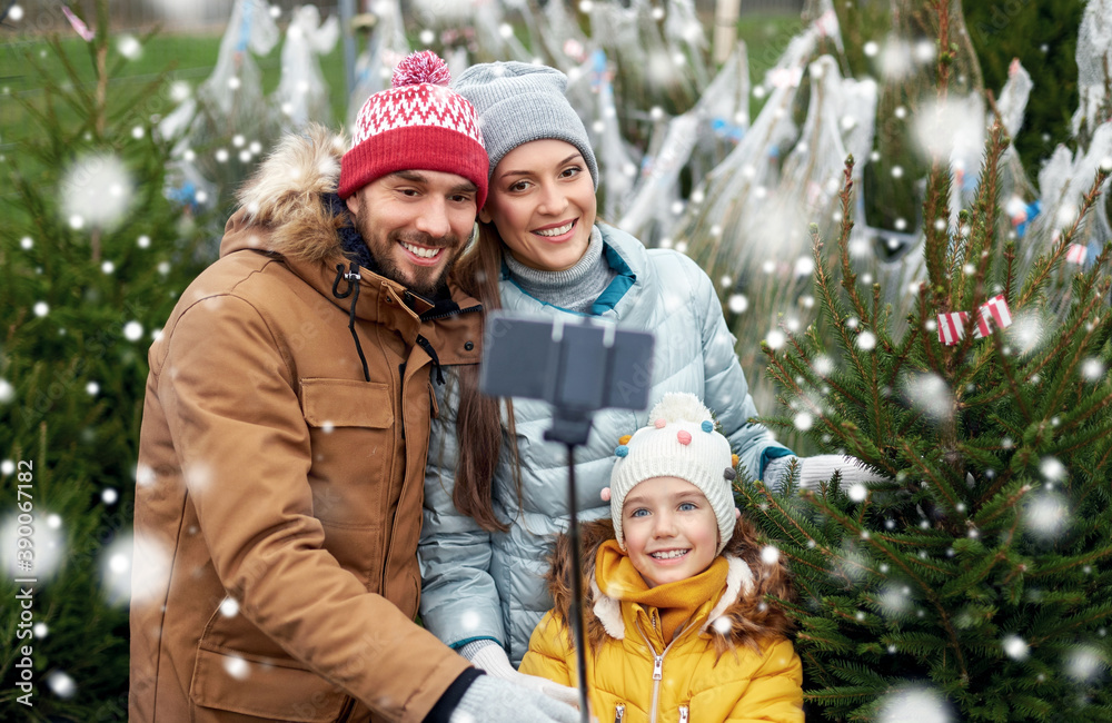 family, winter holidays and people concept - happy mother, father and little daughter buying christmas tree and taking picture with smartphone on selfie stick at street market over snow