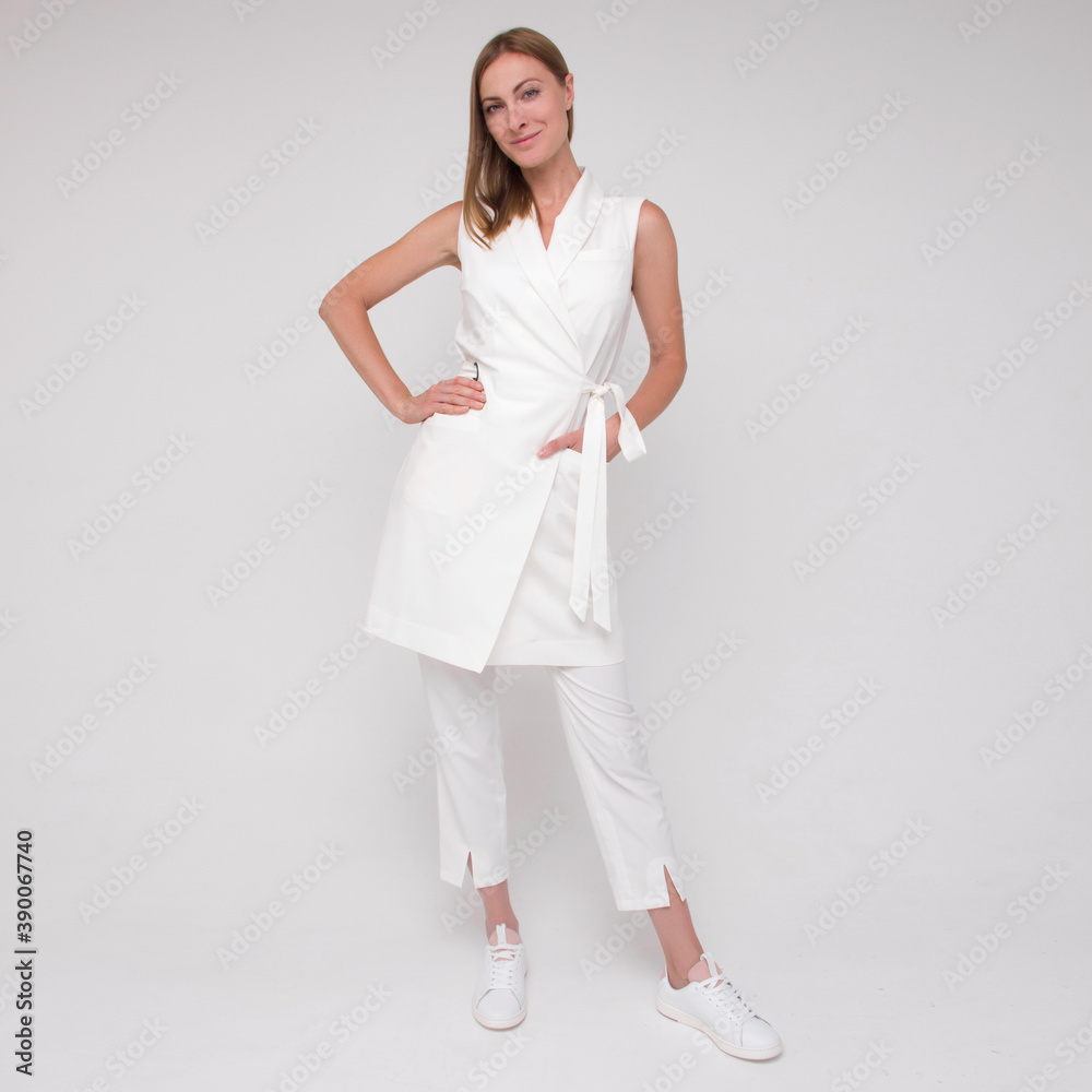 young stylish nurse in white medical costume and white sneakers is standing straight with hands in her pockets and smiling on white wall background. medical concept. free space young stylish nurse in white medical costume and white sneakers is standing straight with hands in her pockets and smiling on white wall background. medical concept. free space