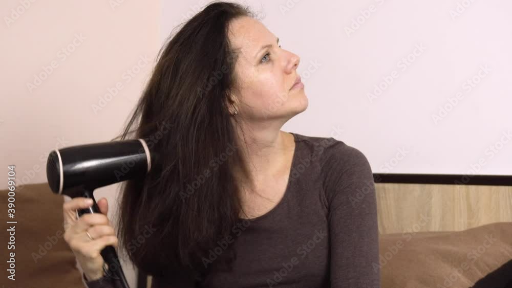 Young beautiful woman dries her hair with a hairdryer on a large bed in her room.Hygiene. Hair care. Hair styling.4k Young beautiful woman dries her hair with a hairdryer on a large bed in her room.Hygiene. Hair care. Hair styling.4k