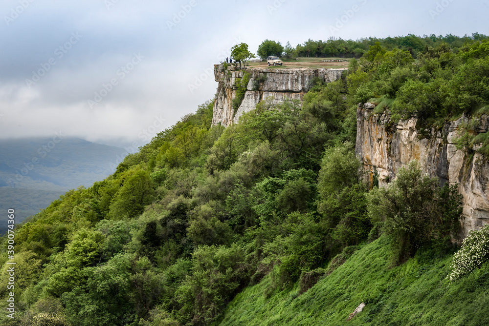 Fototapeta premium BAKHCHISARAI, RUSSIA - 7 MAY, 2019: The rock above the mountain valley with a car on top in the fog on the background of a stormy sky near the town of Bakhchisarai in the Crimean Mountains