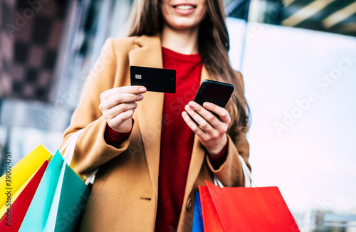 Beautiful trendy young woman with many colorful shopping bags in good mood with smart phone and credit card while walking in the mall during black friday