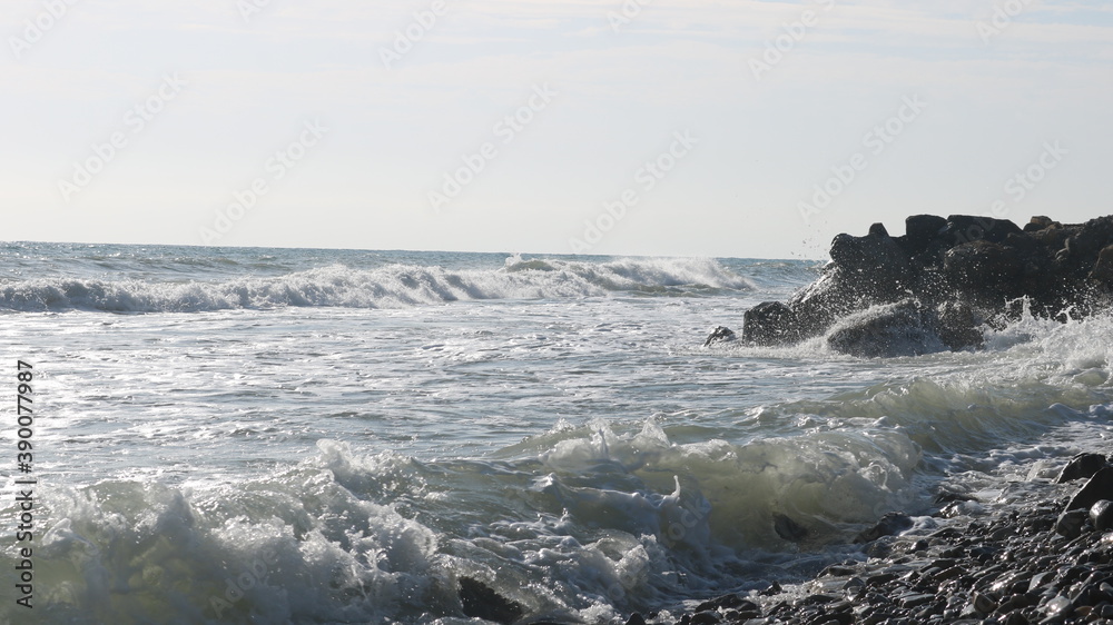 a view of the restless foamy sea with azure greenish waves beating against the black rocks of an empty beach in the sun's rays, giving a metallic reflection of the water surface