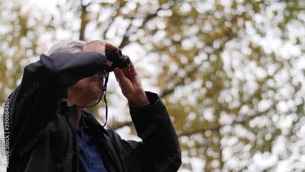 Mature middle-aged white man using binoculars in a scenic forest in late autumn. Handsome guy watches birds outdoors and enjoys his hobby. Beautiful nature background during fall. Happy lifestyle. Mature middle-aged white man using binoculars in a scenic forest in late autumn. Handsome guy watches birds outdoors and enjoys his hobby. Beautiful nature background during fall. Happy lifestyle.