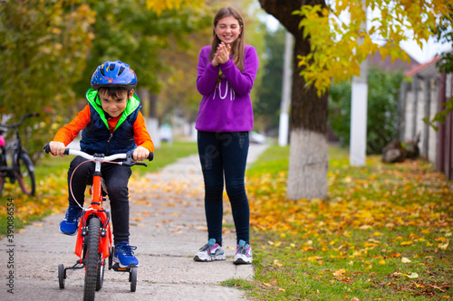Brother and sister in the park. The boy learns to ride a bike alone. The sister in the background rejoices at his success. Photo on the background of autumn.
