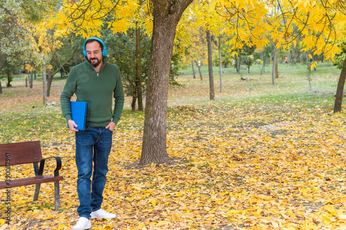 young bearded hipster man standing with blue wireless headphones and laptop under his arm preparing to work in a park in autumn with fallen tree leaves