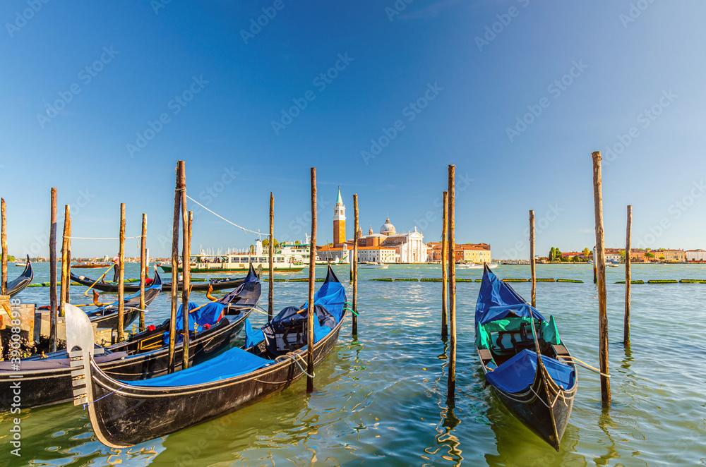 Gondolas moored docked on water in Venice. Gondoliers sailing San Marco basin waterway. San Giorgio Maggiore island with Campanile San Giorgio in Venetian Lagoon, blue clear sky, Veneto Region, Italy