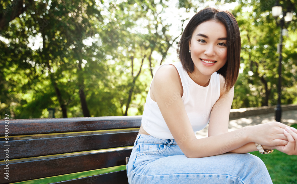 Attractive asian woman relaxing in park. Girl in summer clothes sitting on bench, smiling at camera happy, enjoying sunny day in city Attractive asian woman relaxing in park. Girl in summer clothes sitting on bench, smiling at camera happy, enjoying sunny day in city