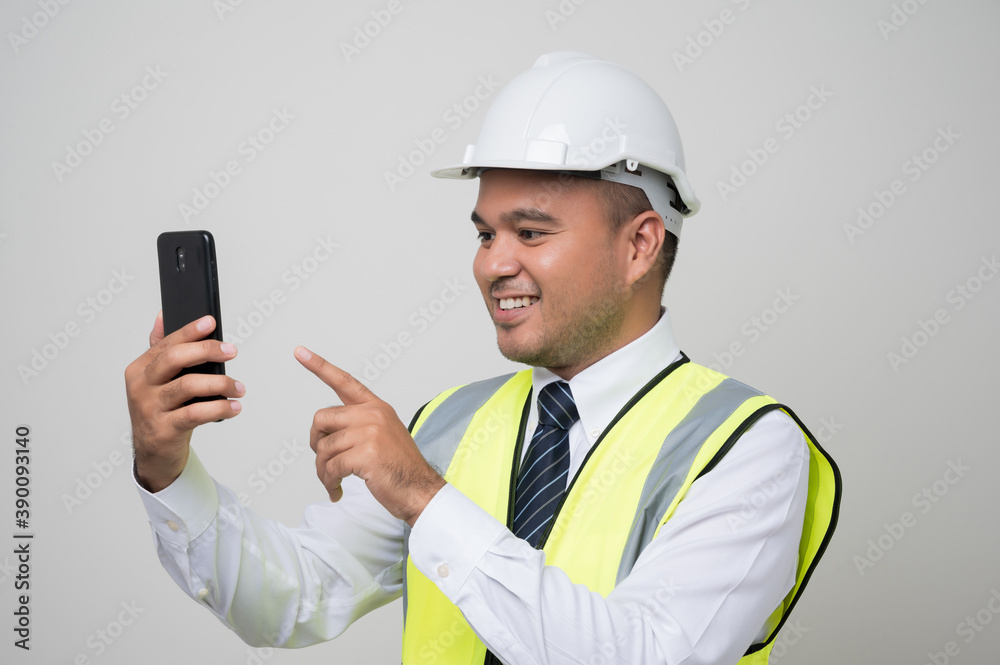 Asian engineer holding mobile phone. Worker wearing hard hat using smartphone standing on isolated white background. Asian engineer holding mobile phone. Worker wearing hard hat using smartphone standing on isolated white background.